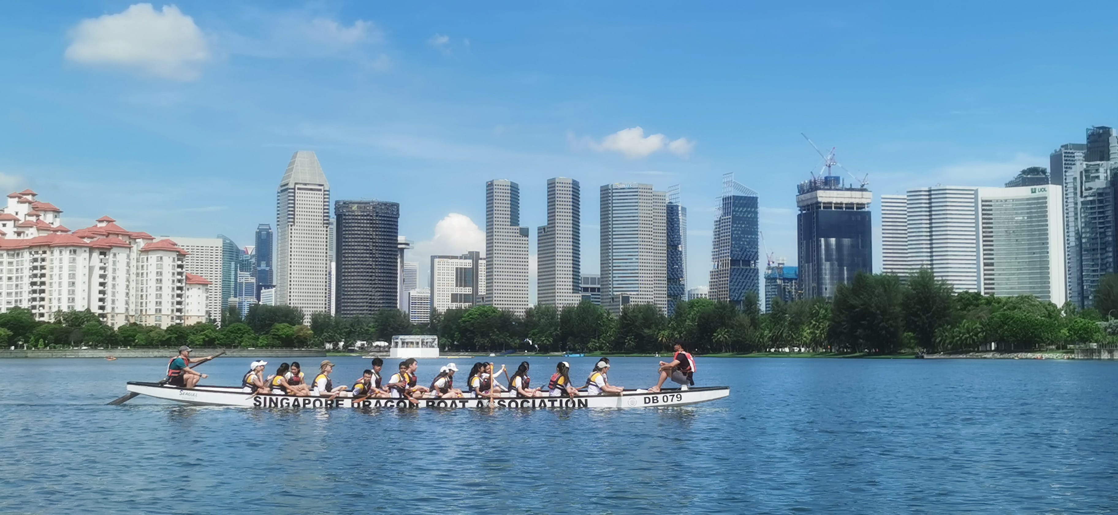 A Singapore Dragon Boat Association team paddles on water with the Singapore city skyline in the background.